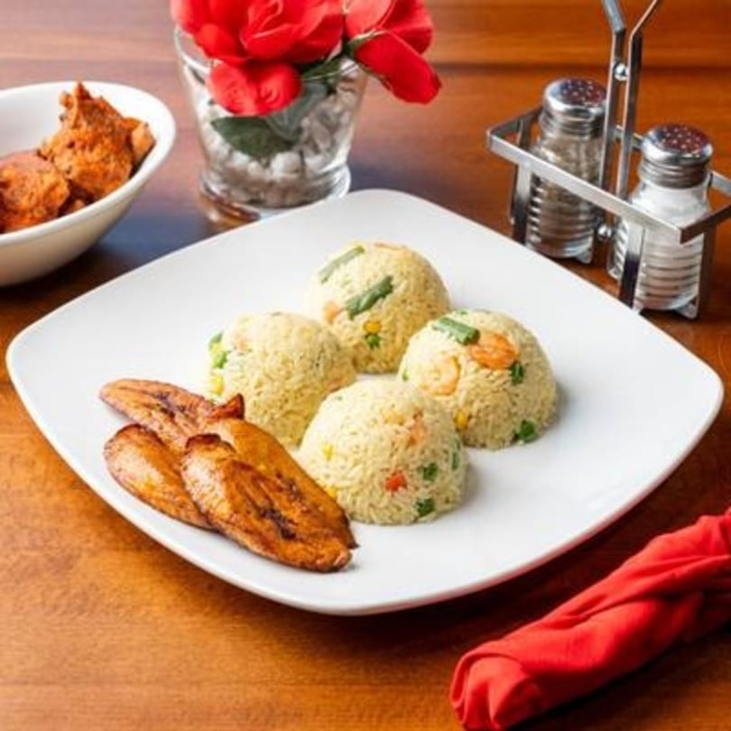 Nigerian fried rice with plantains, a side of chicken, and a red napkin on a wooden table.