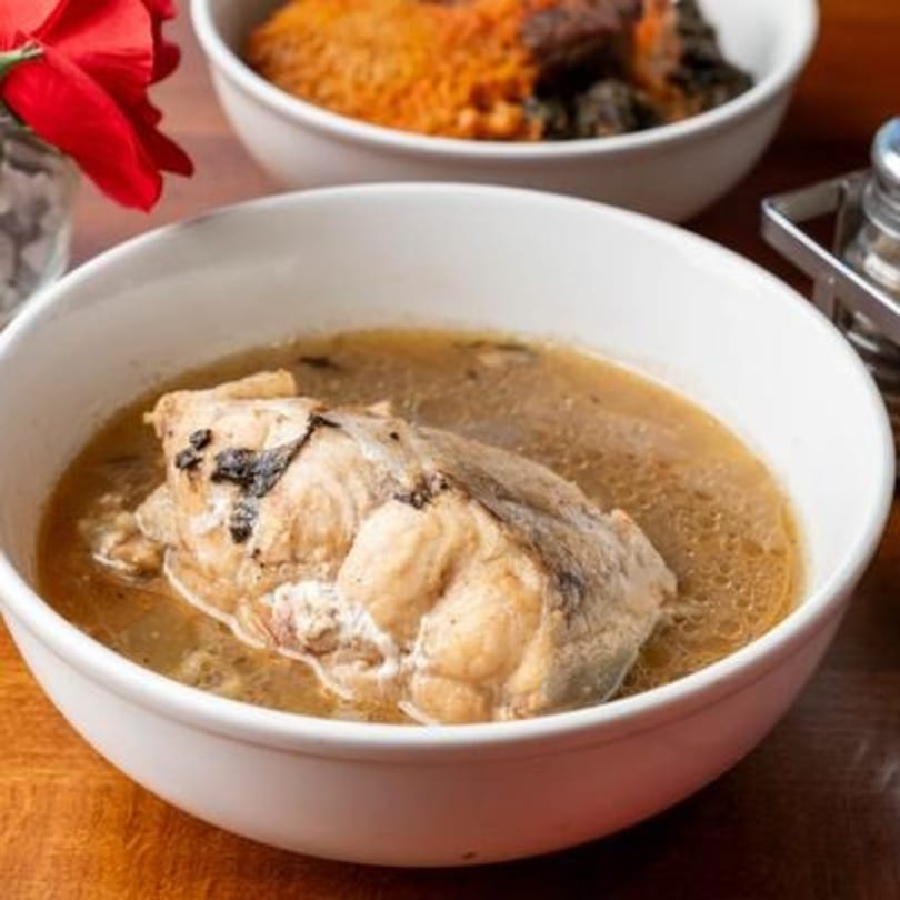 A white bowl of Nigerian fish pepper soup with a large piece of fish in a clear broth.