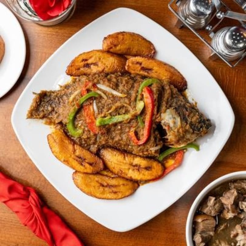 Whole fried fish with plantains and bell peppers on a white plate, with a side of meat stew.