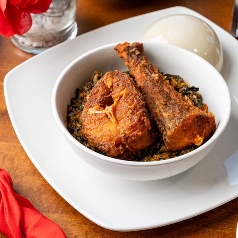 A white bowl of Nigerian stew with fried fish and a side of fufu on a white plate.