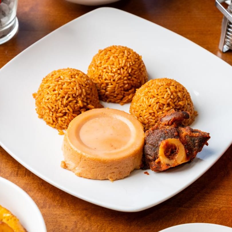 A white plate of Nigerian jollof rice, moin moin, and stewed goat meat.
