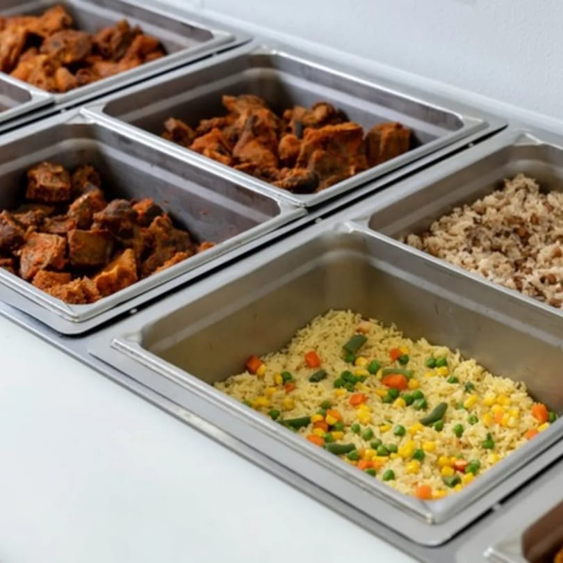 Buffet-style serving dishes filled with Nigerian cuisine, including stewed meat, rice, and mixed vegetables.